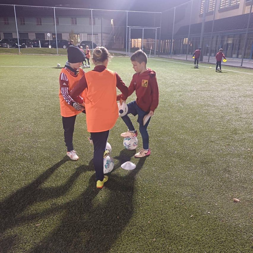 Kinder in orange Westen üben Fußball auf einem Feld mit Kunstrasen bei Nacht, mit anderen im Hintergrund.