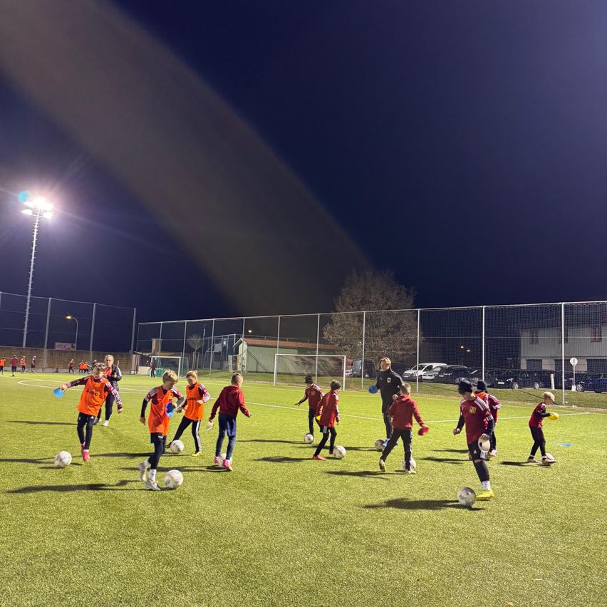 Eine Gruppe von Kindern trainiert nachts auf einem beleuchteten Feld Fußball. Einige tragen orangefarbene Uniformen, andere rote. Sie treten und jagen den Ball.