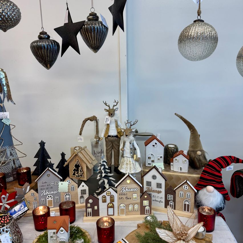 Ein Tisch zeigt verschiedene Weihnachtsdekorationen, darunter Miniaturhäuser, Ornamente und Kerzen, mit einer Rentierfigur und hängenden Ornamenten im Hintergrund.
