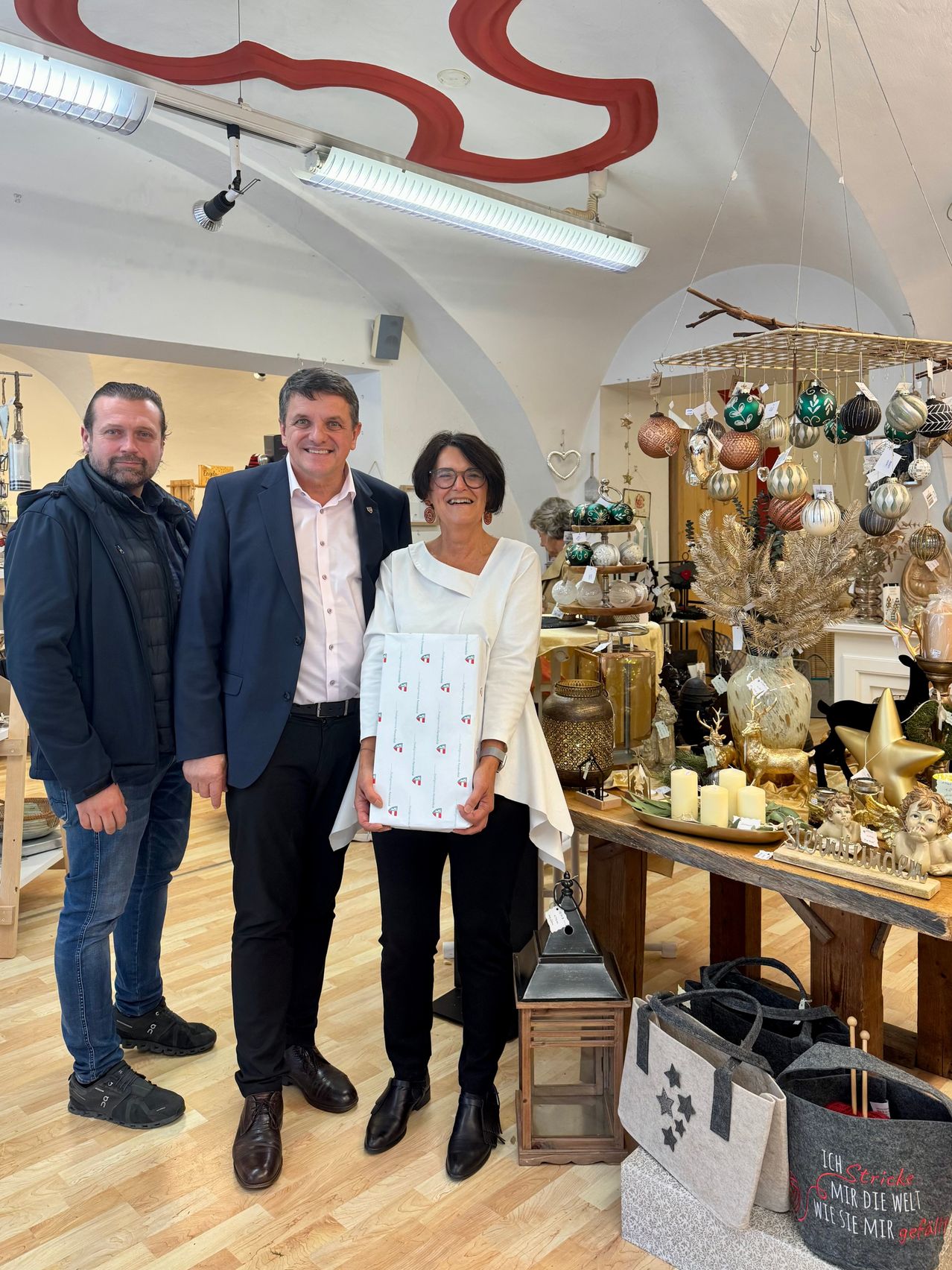 Drei Personen stehen in einem Geschäft und lächeln für ein Foto. Eine Frau hält ein eingewickeltes Geschenk. Hinter ihnen steht ein Tisch mit verschiedenen Ornamenten und einer Lampe.