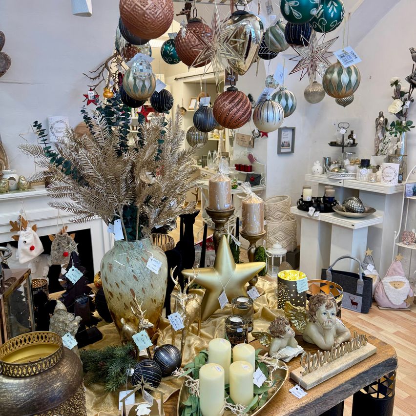 Eine Ausstellung in einem Geschäft mit verschiedenen Ornamenten, Dekorationsartikeln und Kerzen. Eine große Vase mit silbernen Blättern ist prominent ausgestellt, zusammen mit einem goldenen Stern und mehreren dekorativen Engeln. Der Laden hat eine rustikale, aber elegante Ästhetik mit weißen Wänden und Holzböden.