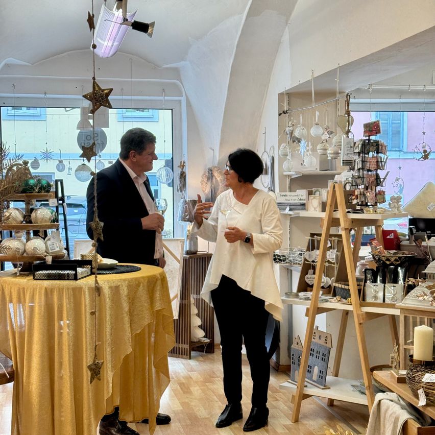 Ein Mann und eine Frau stehen in einem Geschäft und sehen sich an. Der Mann trägt einen Anzug und eine Krawatte, während die Frau eine weiße Bluse und eine Brille trägt. Sie sind umgeben von verschiedenen Dekorationsartikeln und einem Tisch, der mit einem gelben Tuch bedeckt ist.