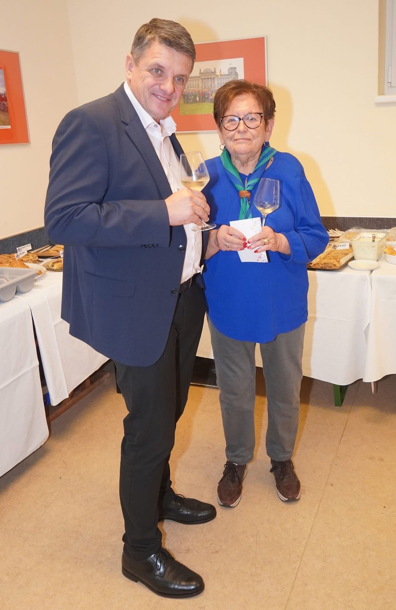 Ein Mann und eine Frau stehen nebeneinander, Weinglas in der Hand, bei einer Veranstaltung. Sie stehen vor einem Tisch mit Speisen und Getränken.
