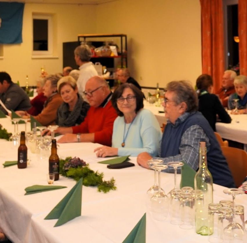 Mehrere ältere Personen sitzen an einem langen Tisch, möglicherweise in einem Speisesaal. Weingläser, Flaschen und grüne Servietten stehen auf dem Tisch. Eine Person in einem weißen Hemd steht hinten.