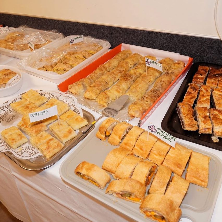Ein Tisch zeigt mehrere Tabletts mit Gebäck. Ein Tablett hat ein Etikett 'Erdappel Strudel'. Ein weiteres Tablett ist mit 'Erdappel' beschriftet. Eine Schüssel hält Gebäck. Ein orangefarbenes Tablett mit Gebäck hat eine weiße Karte mit der Aufschrift 'Erdappel'.