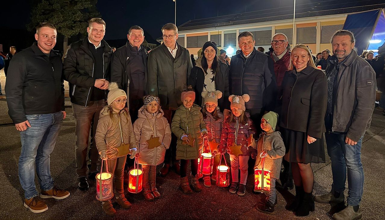 Eine Gruppe von Erwachsenen und Kindern steht bei Nacht zusammen und hält Laternen. Sie sind warm für das kalte Wetter gekleidet. Hinter ihnen steht ein Gebäude mit einer Straßenlaterne.