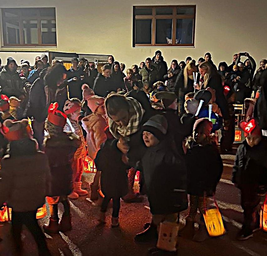 Eine Menschenmenge, darunter Kinder in Kostümen und Mützen, versammelt sich bei Nacht im Freien. Einige halten Laternen, andere haben Kameras. Ein Gebäude mit Glasfenstern steht hinter ihnen.