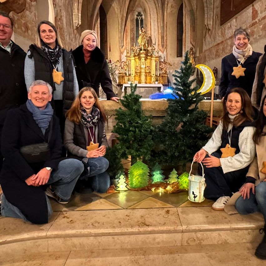 Eine Gruppe von Menschen posiert für ein Foto in einer Kirche mit dekorativen Elementen. Sie tragen Winterkleidung und lächeln, mit Bäumen und einem goldenen Altar im Hintergrund.
