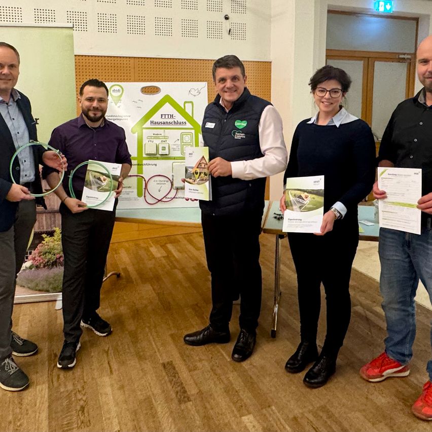 Fünf Personen stehen in einem Raum, halten Urkunden in der Hand und lächeln für die Kamera. Hinter ihnen befindet sich ein Poster mit einem Haussymbol und der Aufschrift 'FTTH-Hausanschluss'.