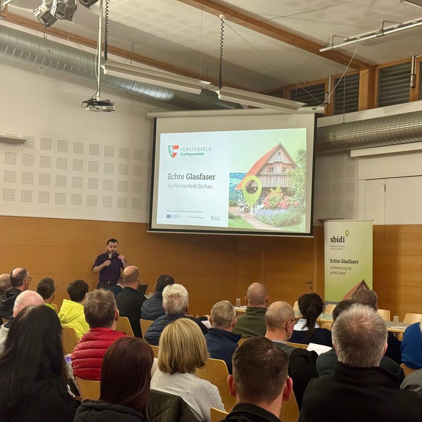 Ein Mann steht vor einer Projektionsleinwand und hält eine Präsentation. Ein Banner steht rechts neben dem Mann. Das Publikum sitzt in Stühlen vor ihm.