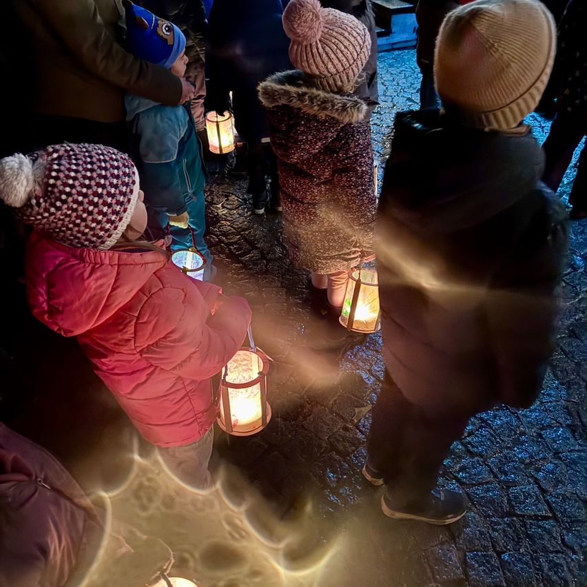 Kinder in Wintermänteln und Mützen halten brennende Laternen in einem schwach beleuchteten Außenbereich.