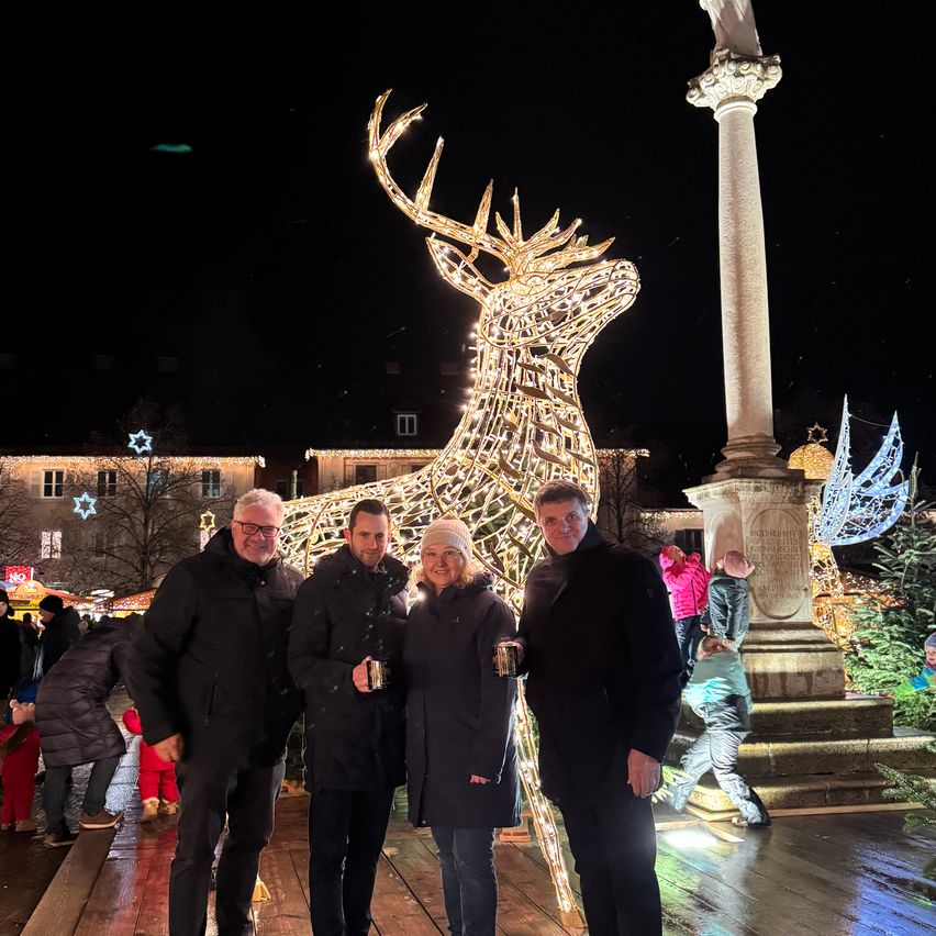 Vier Personen stehen vor einer beleuchteten Rehenskulptur und lächeln für ein Foto. Dahinter ist eine festlich dekorierte Straße zu sehen. Im Hintergrund steht eine hohe Steinsäule und verschiedene Weihnachtsbäume.