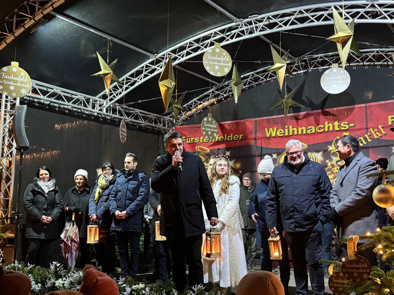 Eine Gruppe von Menschen auf einer Bühne bei einem Weihnachtsevent, ein Mann spricht in ein Mikrofon, während andere Laternen halten.