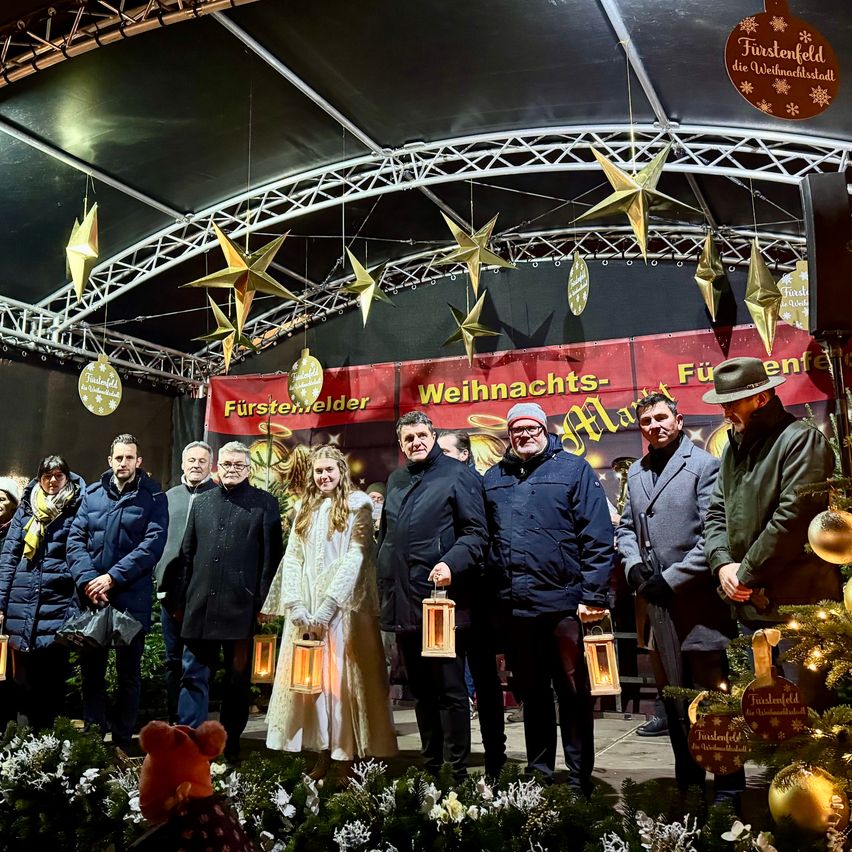 Eine Gruppe von Menschen in Winterkleidung steht vor einem geschmückten Weihnachtsmarkt. Der Markt ist mit einem Vordach bedeckt, das mit Sternenverzierungen geschmückt ist. Ein Weihnachtsbaum und ein roter Banner sind in der Szene zu sehen.