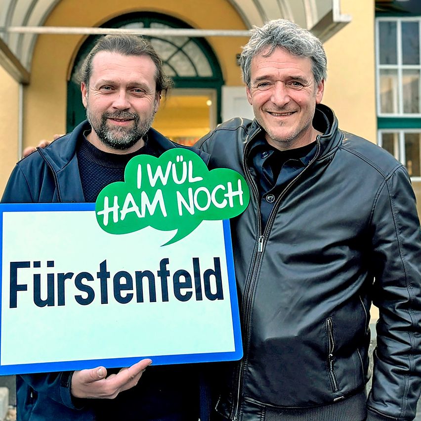 Two men stand outside near a building, one holds a sign that says 'Fürstenfeld' and has a speech bubble with 'I wül HAM noch'.