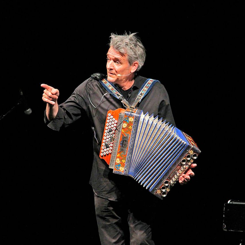 A man with gray hair is standing on a stage holding an accordion, he is pointing at something.