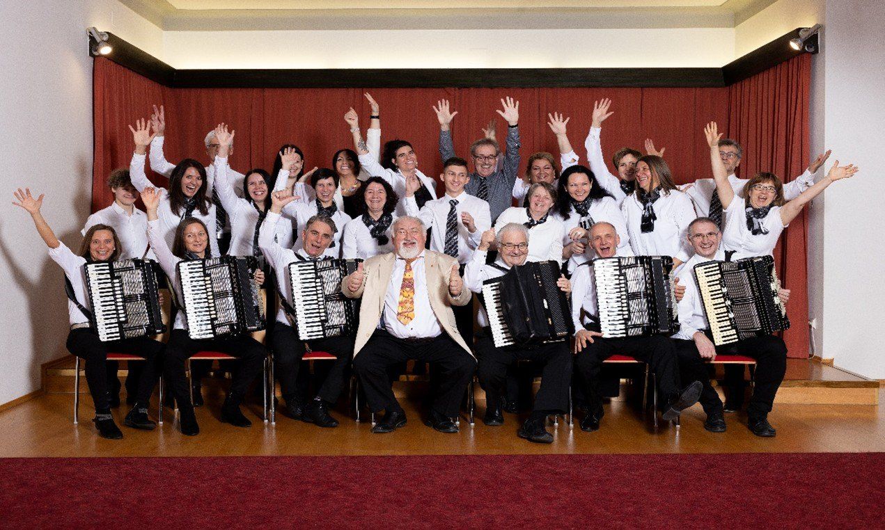 A group of musicians in white shirts and ties, seated and standing, holding accordions, posing for a photo in front of a red curtain.