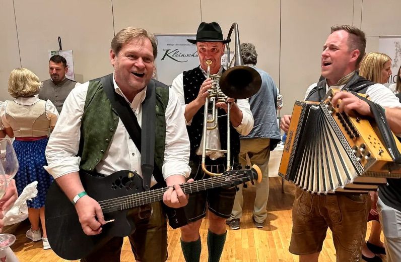 A group of men in traditional German attire play instruments at an event. One man plays a trombone, while others play guitar and accordion. They are smiling and appear to be enjoying themselves.