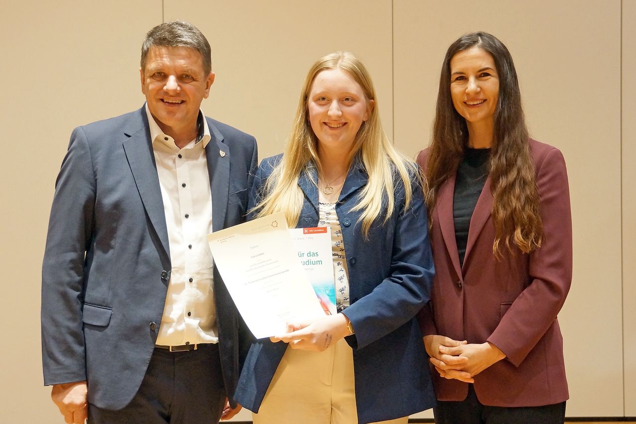Drei Personen stehen in einem Raum und lächeln. Die Frau hält ein Zertifikat und ein Buch, während der Mann links ein weißes Hemd trägt und der andere Mann rechts einen roten Anzug.