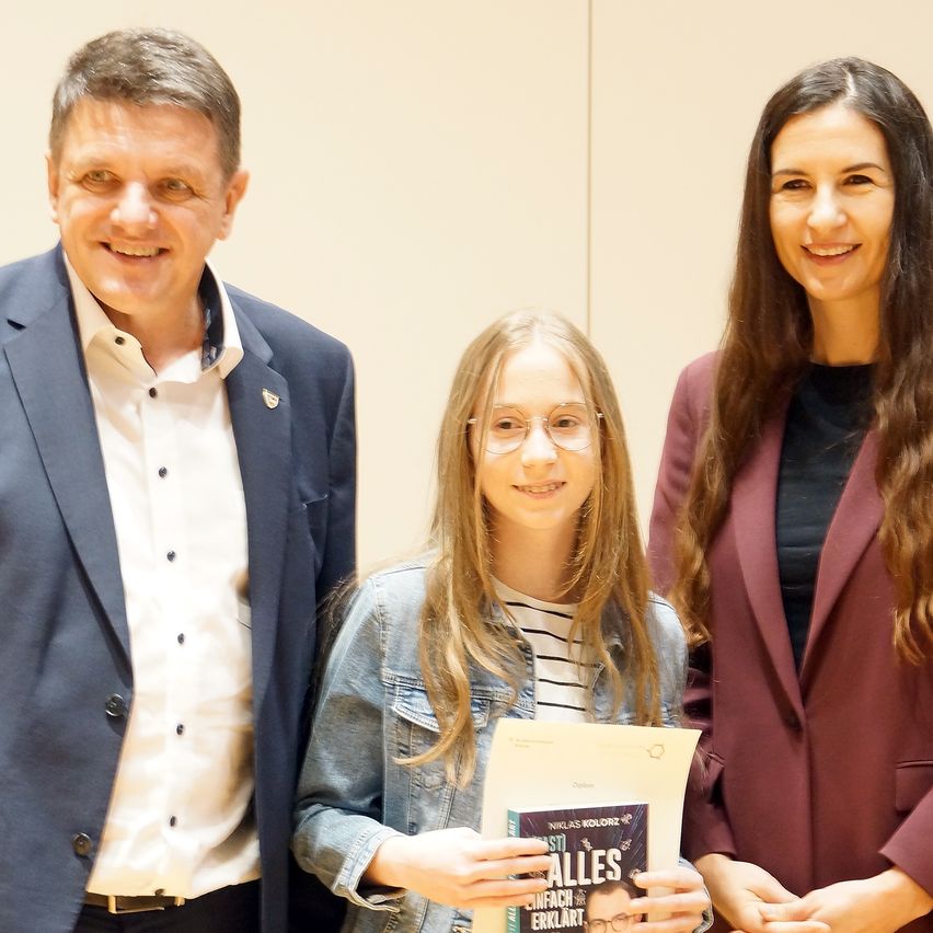 Drei Menschen lächeln und posieren für ein Foto. Das Mädchen hält ein Buch mit dem Titel Alles Anfach Erklärt. Der Mann trägt einen Anzug. Die Frau trägt einen kastanienbraunen Blazer.