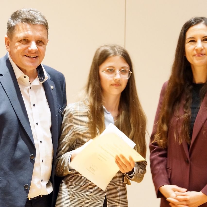 Drei Personen stehen in einem Raum; ein Mann und zwei Frauen, lächelnd. Der Mann trägt einen dunkelblauen Anzug und ein weißes Hemd. Die Frau auf der linken Seite hält eine Mappe und trägt eine Brille. Die Frau auf der rechten Seite hat lange Haare.