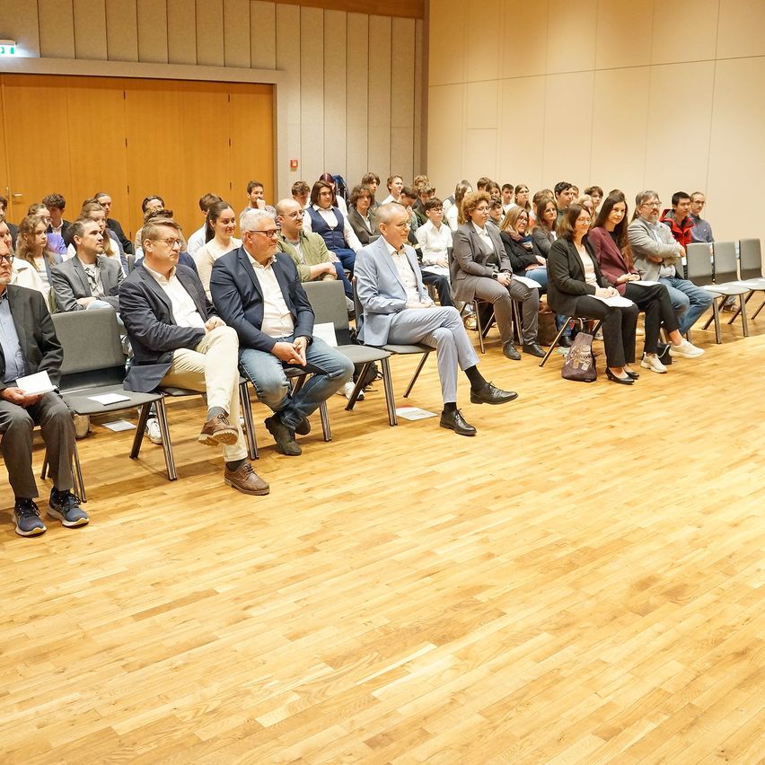 Ein Konferenzraum mit Holzboden hat Reihen von Stühlen. Die Teilnehmer sitzen und einige halten Papiere. Ein paar Leute stehen in der Nähe einer Holzwand.