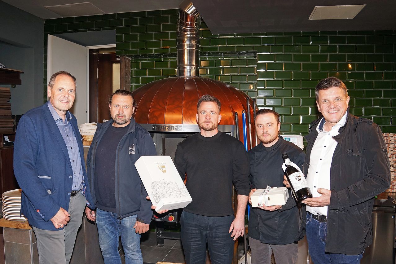 Fünf Männer posieren für ein Foto vor einer grün gefliesten Wand, jeder hält ein Geschenkkästchen und eine Flasche Wein in der Hand. Sie stehen vor einem großen kupfernen Ofen.