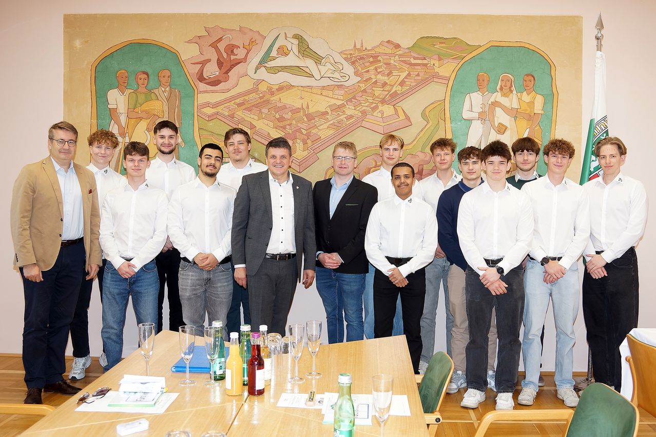 Eine Gruppe von Menschen in weißen Hemden steht in einem Raum mit einem Wandbild. Ein Tisch mit Weingläsern und Flaschen steht vor ihnen.