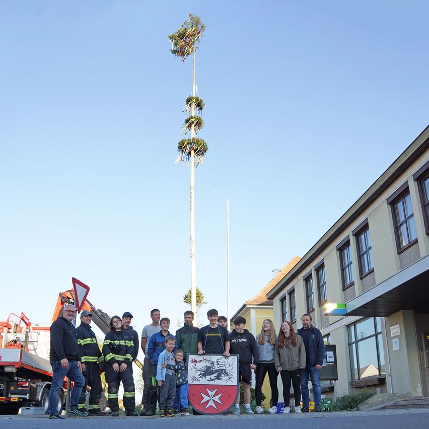 Eine Gruppe von Menschen posiert für ein Foto vor einem hohen Mast mit Dekorationen. Hinter ihnen steht ein Gebäude mit einem roten Schild. Links steht ein Feuerwehrauto.