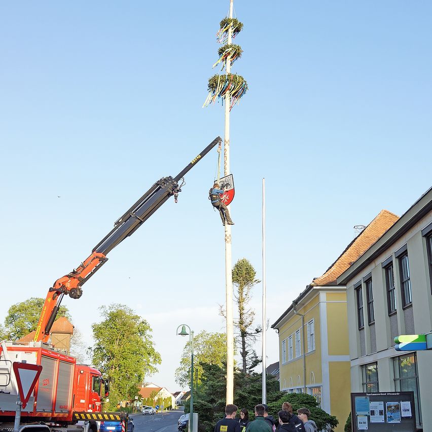 Ein Kran hebt einen Mann zur Spitze eines hohen Mastes, wo er die Girlanden entfernt. Leute beobachten dies auf der Straße.