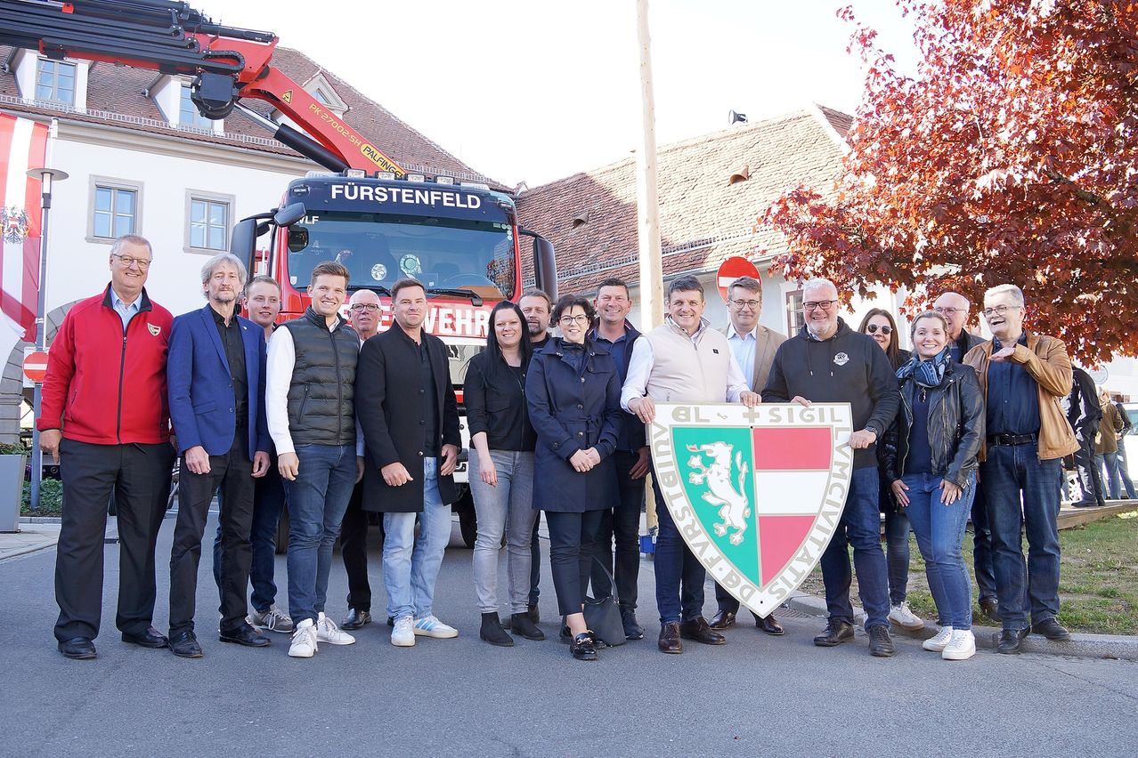 Eine Gruppe von Menschen posiert für ein Foto mit einem Schild und einem Feuerwehrauto namens Furstenfeld im Hintergrund.