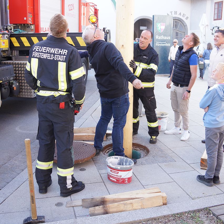 Feuerwehrleute aus Furstenfeld-Stadt arbeiten an einem Holzpfosten, eine Farbeimer daneben. Zuschauer beobachten.