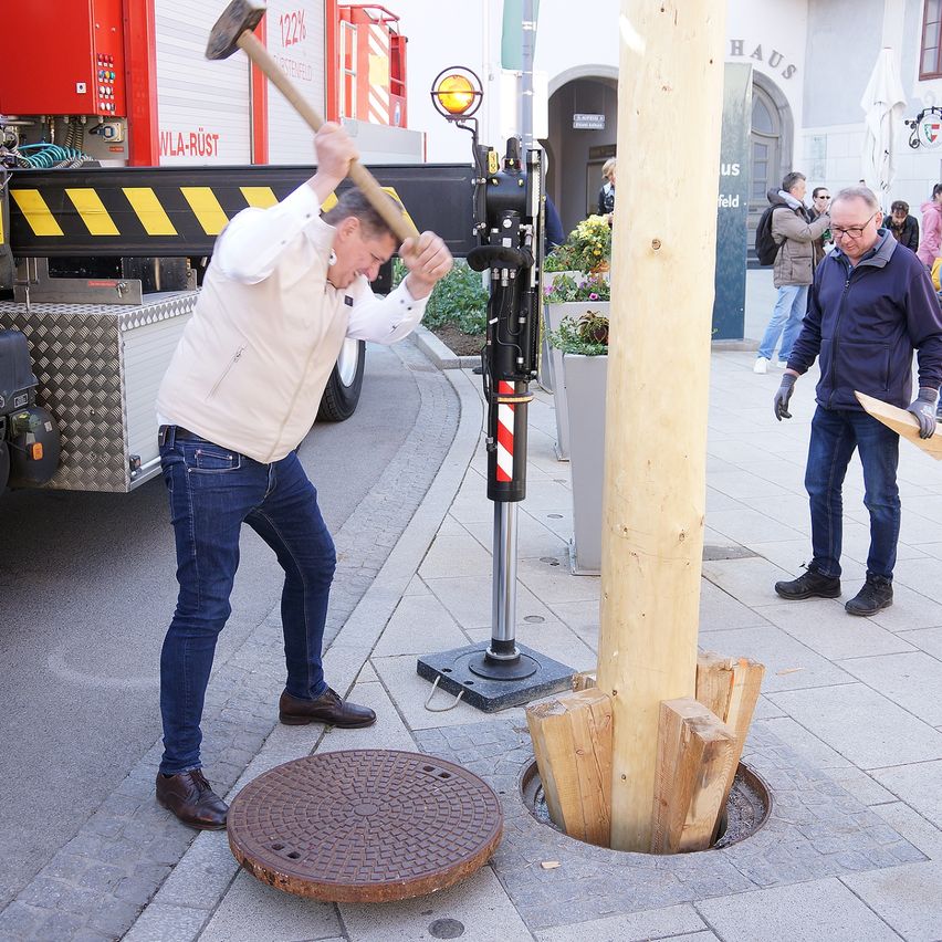 Ein Mann schlägt mit einem Vorschlaghammer auf einen Holzpfahl, während ein anderer Mann zusieht. Hinter ihnen ist ein Feuerwehrauto geparkt und mehrere Menschen gehen auf dem Bürgersteig.