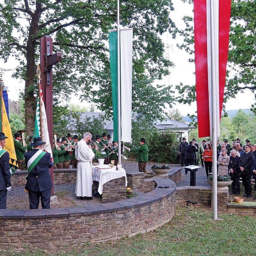 A priest is conducting a ceremony in a garden area with flags and a crowd of people.