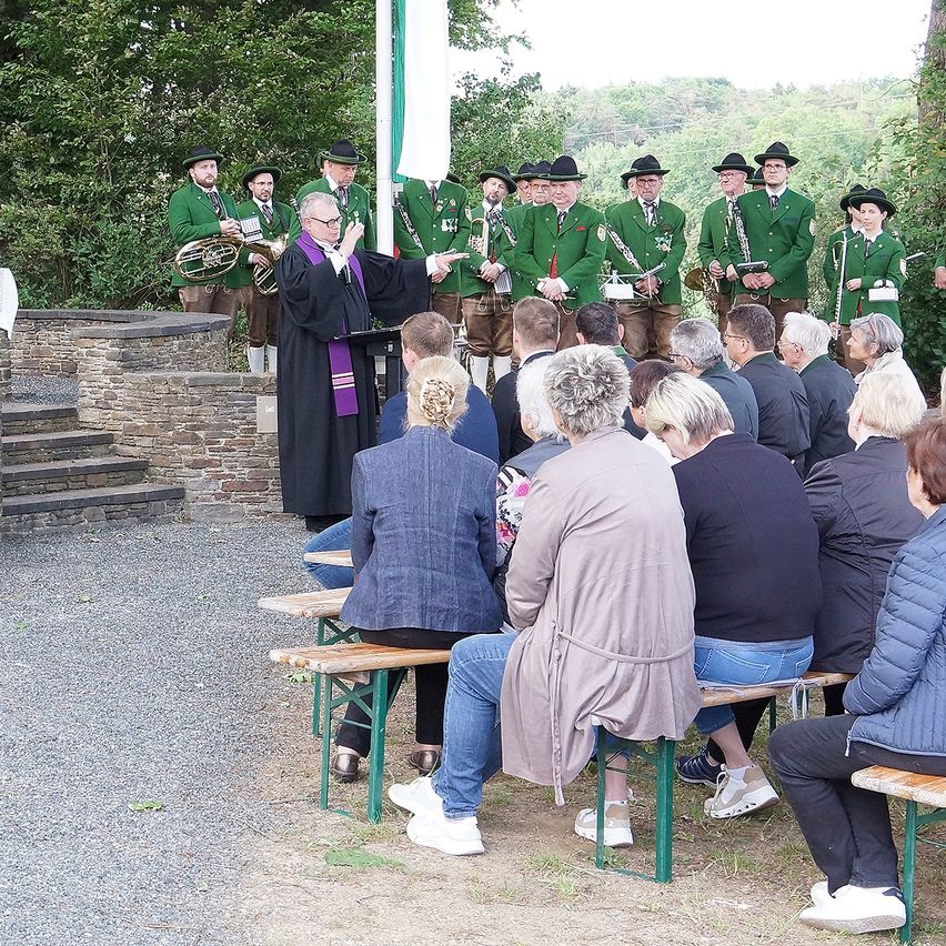 A group of people dressed in green jackets and hats are standing and performing with musical instruments. An elderly man in a black robe is speaking to them. Behind them, several people are sitting on benches watching.