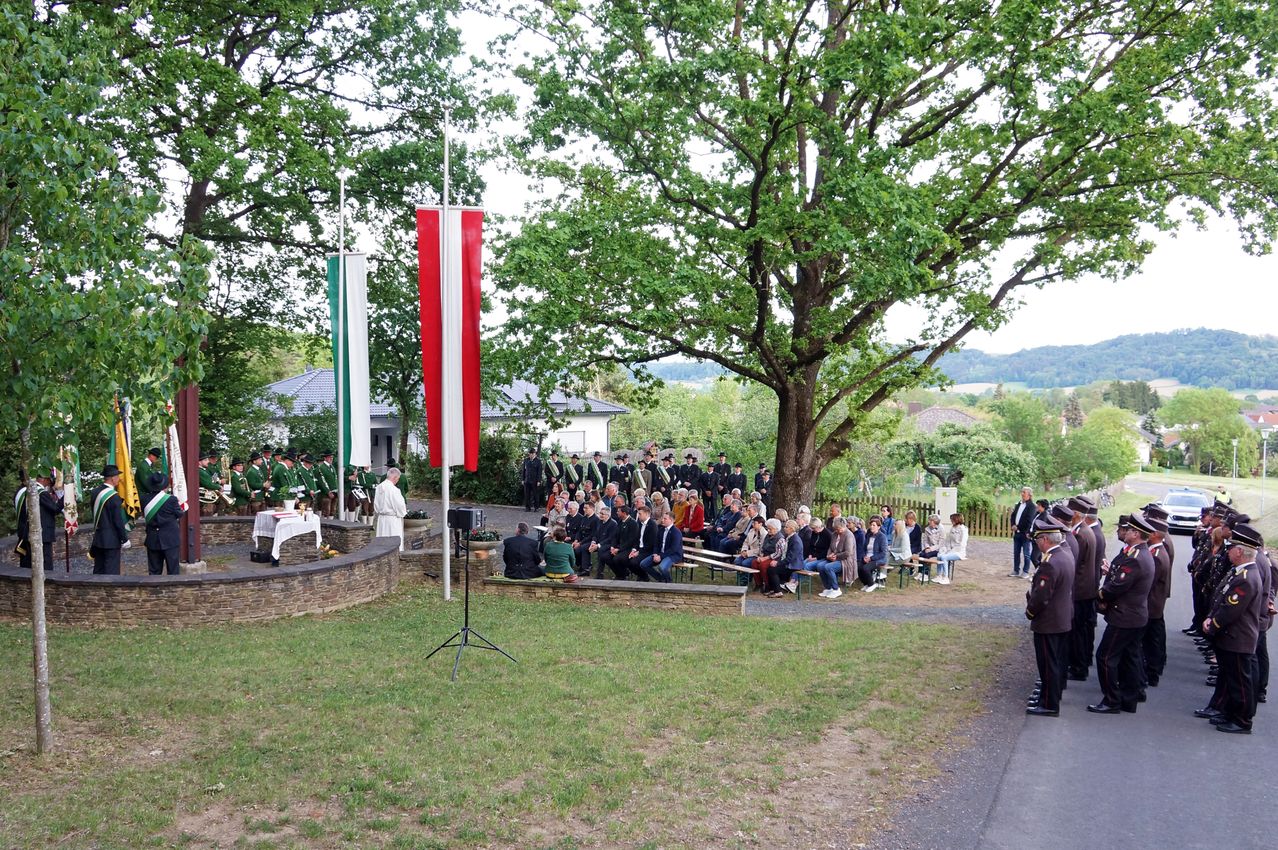 A gathering of people in formal attire is seated under a tree. Flags are hoisted on poles in the background. A man stands on a podium with a microphone.