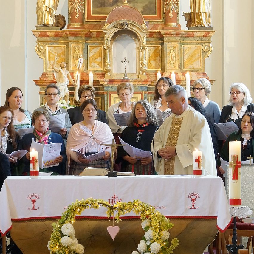 A group of people stand at an altar, possibly during a religious ceremony. They hold papers, and candles are on the altar. A priest stands at the center, looking down.