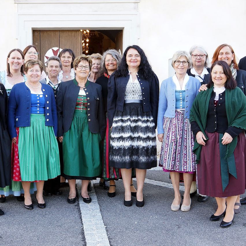 A group of women dressed in traditional clothing stands in front of a white building, smiling for a photo.