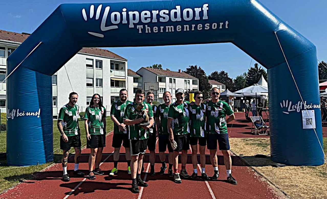 A group of men and women in green jerseys stand under a blue arch with 'Loipersdor Thermenresort' written on it. They are on a red track, holding a trophy and posing for a photo.
