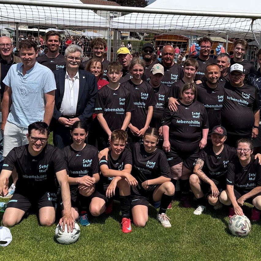 A group of people in black jerseys with Super Kraft written on them pose for a photo in front of a soccer goal with tents in the background.