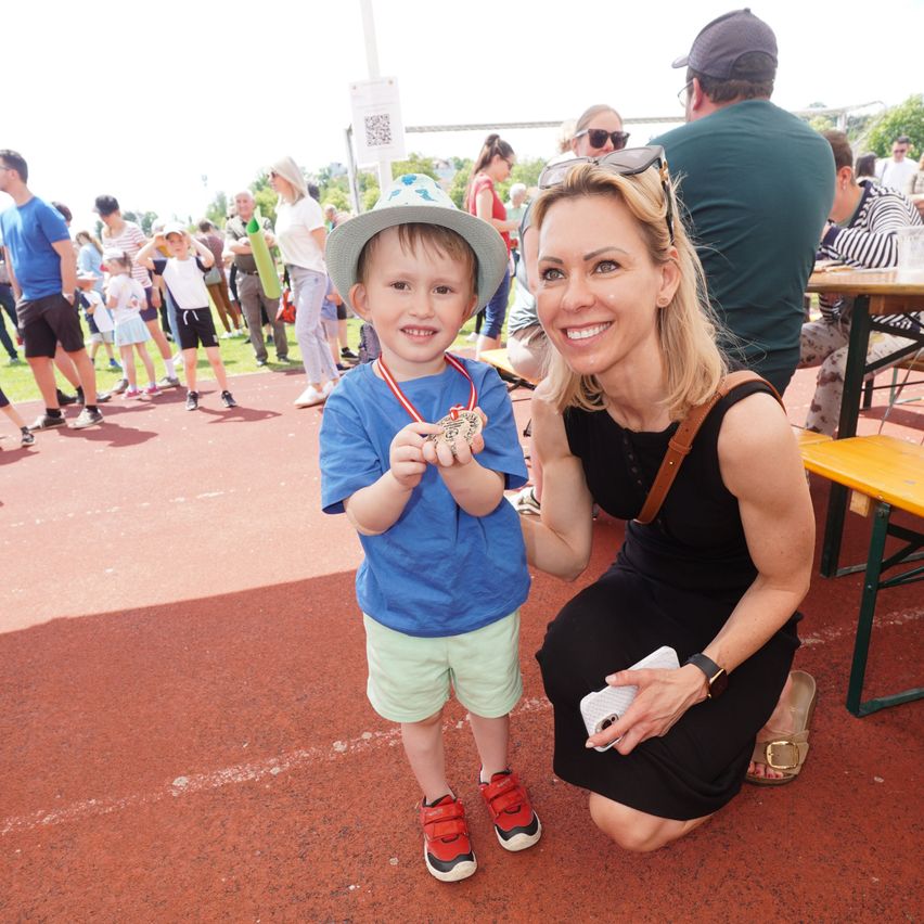 A young boy with a medal and a woman smiling at a track event, with other people walking and sitting in the background.