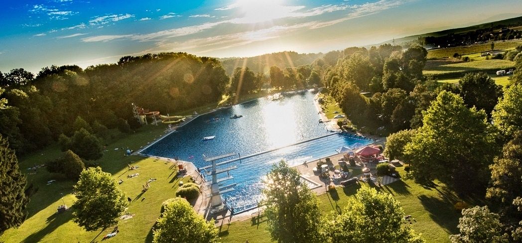 Eine Luftaufnahme eines großen Schwimmbeckens, umgeben von viel Grün, mit Sonnenbadenden und einer strahlenden Sonne.