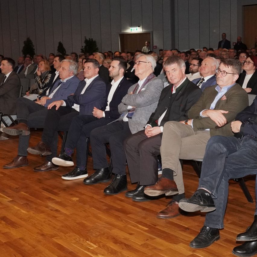Ein überfüllter Raum mit formell gekleideten Männern und Frauen, die in Reihen auf Stühlen sitzen und aufmerksam zuhören, mit einer Bühne im Hintergrund.