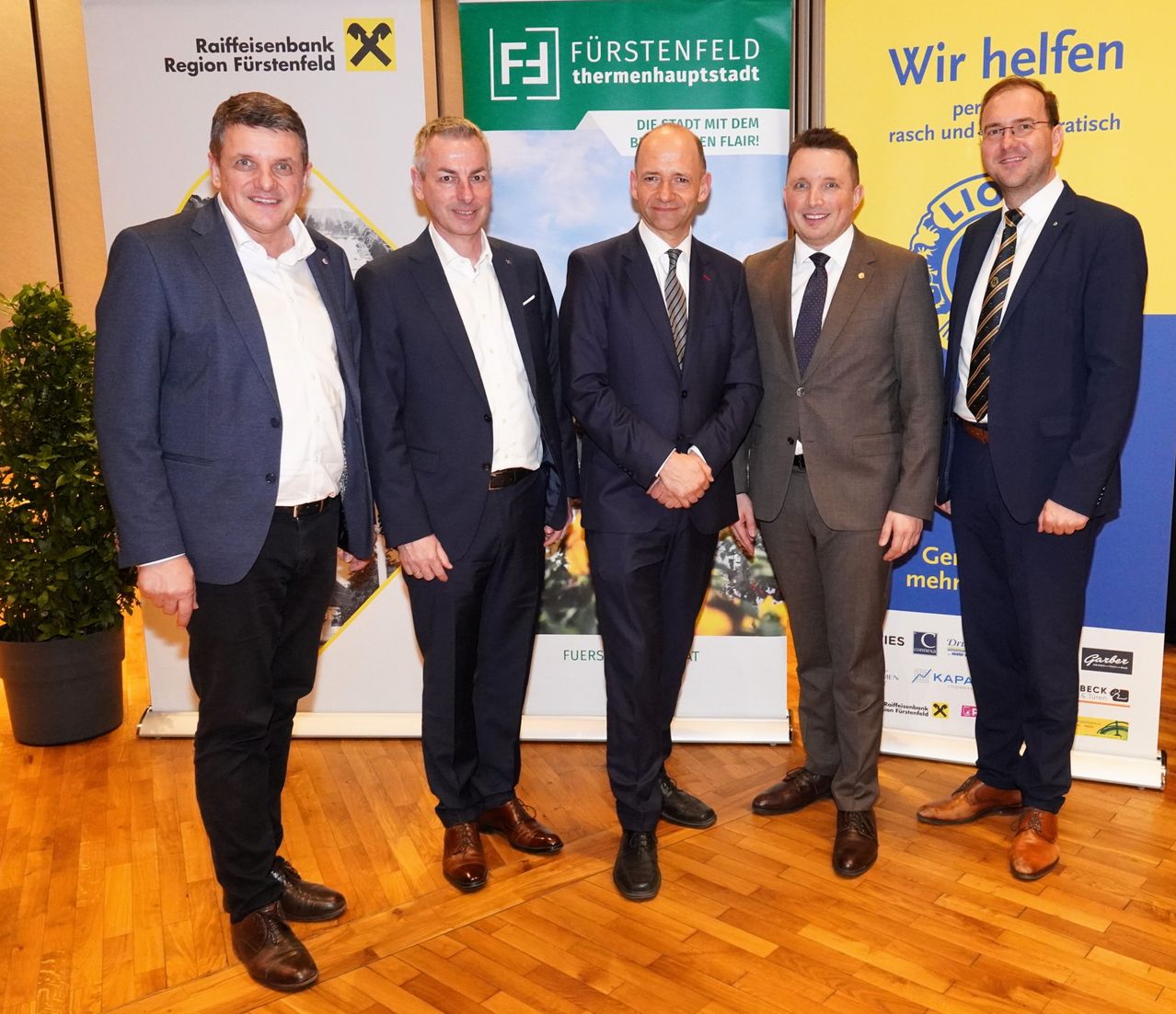 Fünf Männer in formeller Kleidung stehen vor Bannern und einem Holzboden. Zwei Banner an den Seiten zeigen den Text Raiffeisenbank und Furstenfeld.