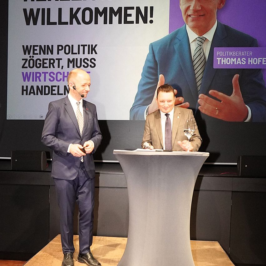 Zwei Männer in Anzügen an einem Rednerpult mit einer großen Leinwand dahinter. Die Leinwand zeigt eine politische Botschaft. Ein Mann spricht, während der andere zuhört.