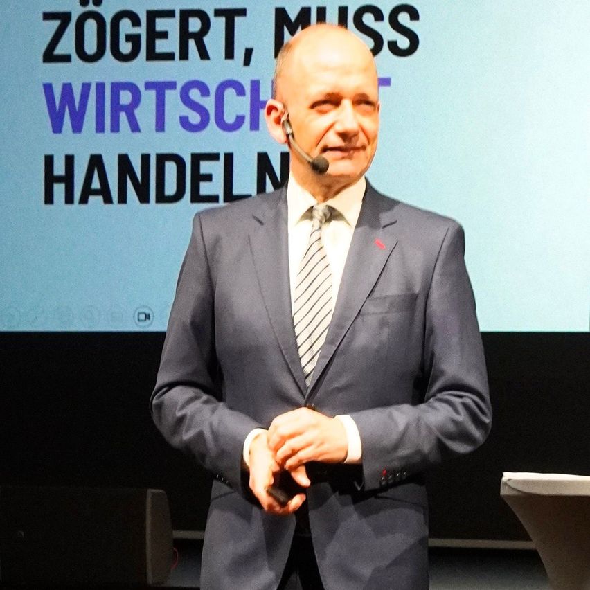 Ein Mann in Anzug und Krawatte steht auf der Bühne, hält ein Mikrofon in der Hand und spricht vor einem Publikum. Hinter ihm zeigt eine große Leinwand die Worte 'Zog, muss wirtschaften'.