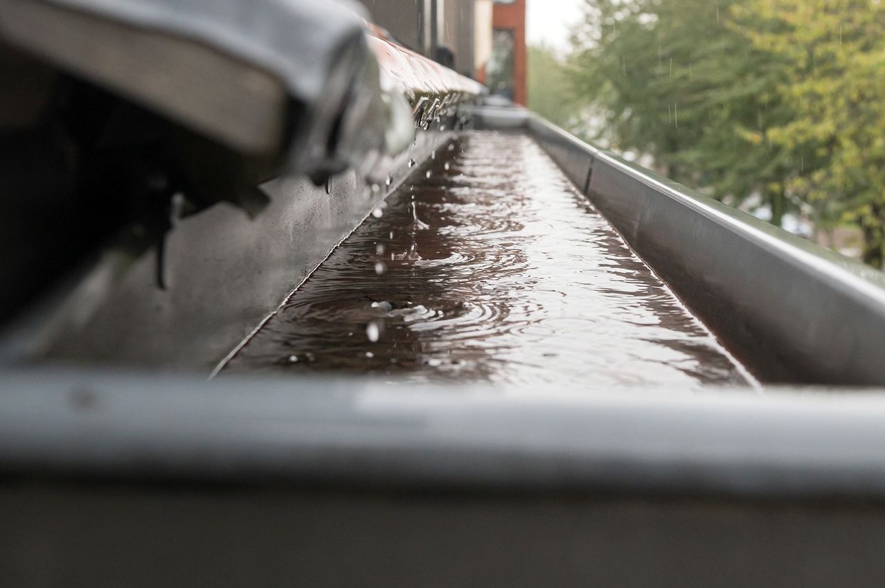 Nahaufnahme von Regenwasser, das während eines Regentages durch eine Dachrinne fließt. Bäume im Hintergrund deuten auf eine Außenumgebung hin.