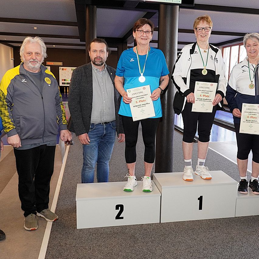 Vier Personen stehen auf einem Podest. Die beiden Frauen in der Mitte halten Urkunden und Medaillen. Der Mann links trägt eine Jacke und der Mann rechts trägt eine Brille.