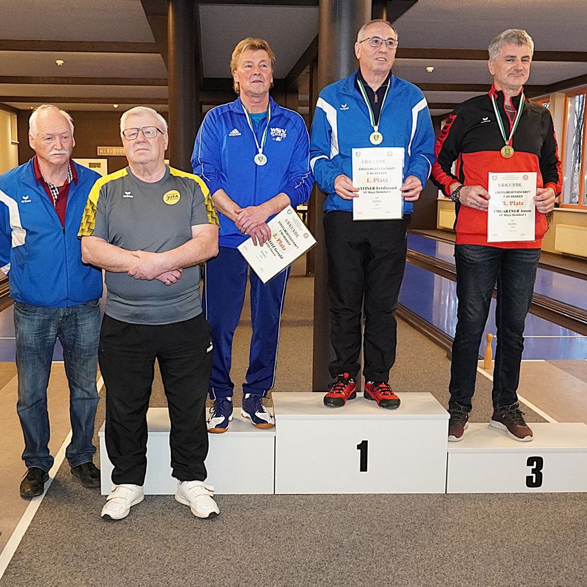 Fünf Männer stehen auf einem Podium mit Medaillen und Urkunden. Ein Mann hält ein Zertifikat, während ein anderer einen Pokal in der Hand hat. Sie tragen alle Jacken und Brillen. Der Boden ist mit Teppich ausgelegt, und eine Bowlingkegel ist im Hintergrund zu sehen.