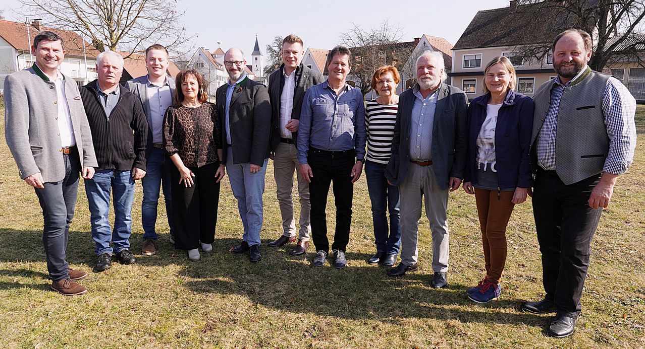 Eine Gruppe von Personen steht auf einem Rasenfeld mit Häusern im Hintergrund. Sie sind alle formell gekleidet. Die Männer tragen Anzüge und Lederschuhe, während die Frauen Kleider und Schuhe tragen. Sie scheinen für ein Foto zu posieren.